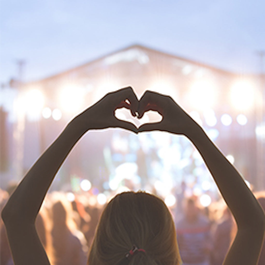 Frau auf einem Konzert. Darstellung von hinten, die ihre Arme über den Kopf hält und mit den Händen ein Herz formt